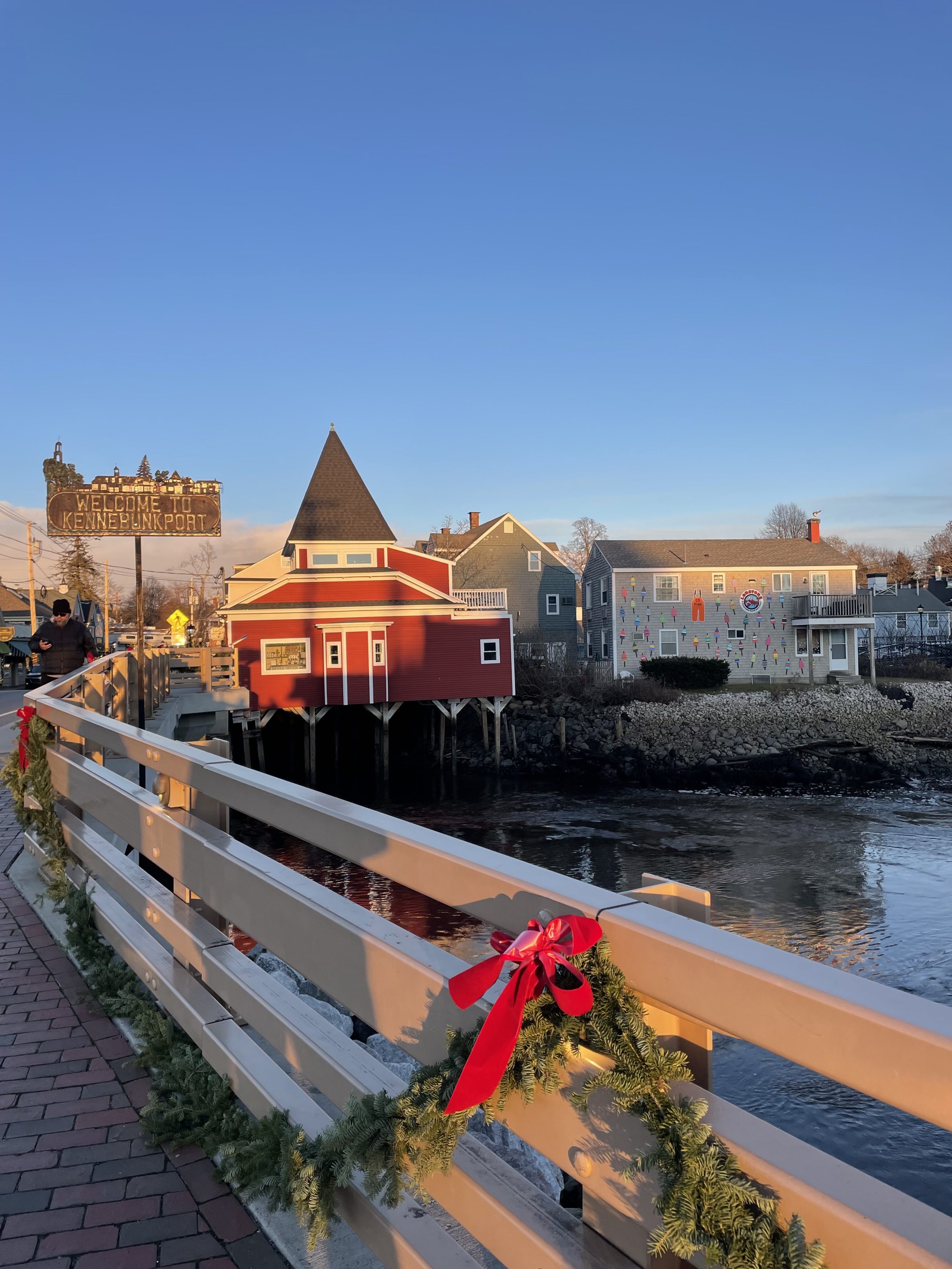 A building with a red bow located on a bridge over water in Kennebunkport. The surroundings include outdoor landscape with a lake and a city in the background.