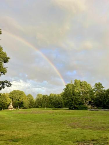 Lush green lawn with trees and a monument in the background, under a cloudy sky adorned with a vibrant rainbow.
