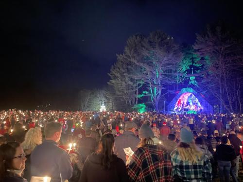 Nighttime scene of carolers gathered in front of a grotto nestled among trees illuminated with colorful lights. 