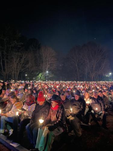 Nighttime scene of a lawn filled with hundreds of carolers, each holding a glowing candle.